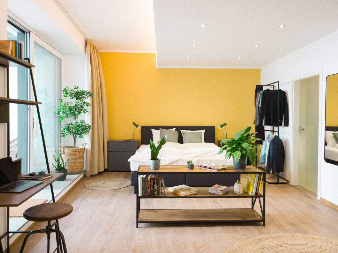 Apartment mit sonnengelber Wand, Bett, Pflanzen und großen Fenstern