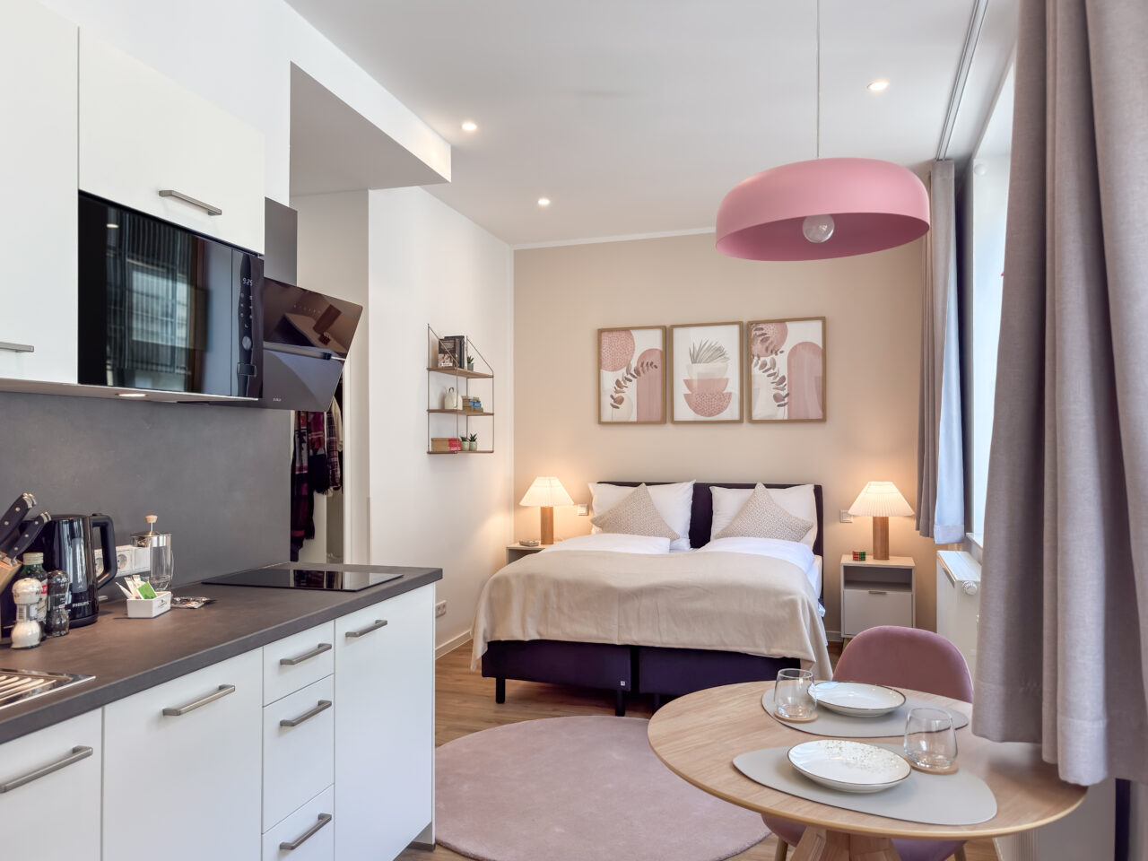 Weiße Küchenzeile mit rosa Pendelleuchte und Bett im Hintergrund