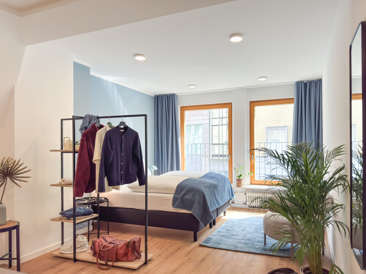 Bett vor hellblauer Wand mit Kleiderstange und großer Pflanze